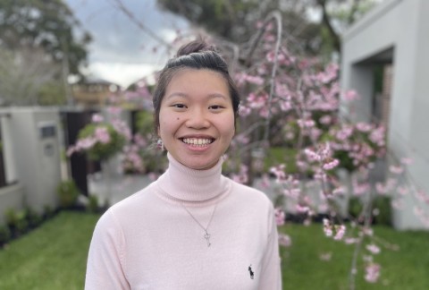 Jeanette smiles while standing outside, there is a tree with pink blossoms behind her in full bloom.